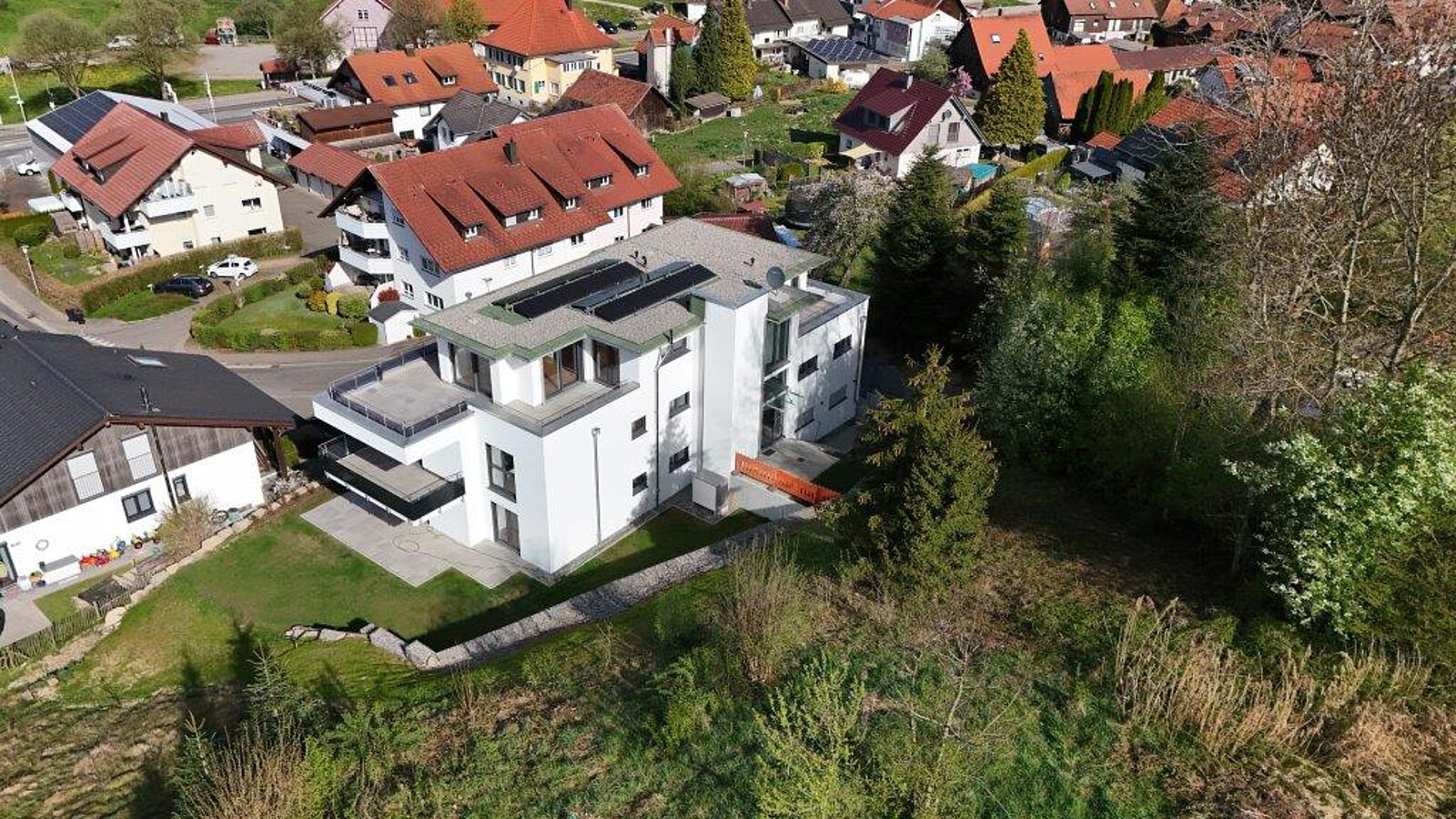 Luftaufnahme von Neuravensburg mit der historischen Burgruine auf einem bewaldeten Hügel im Hintergrund und einem modernen Wohnhaus am Ortsrand.