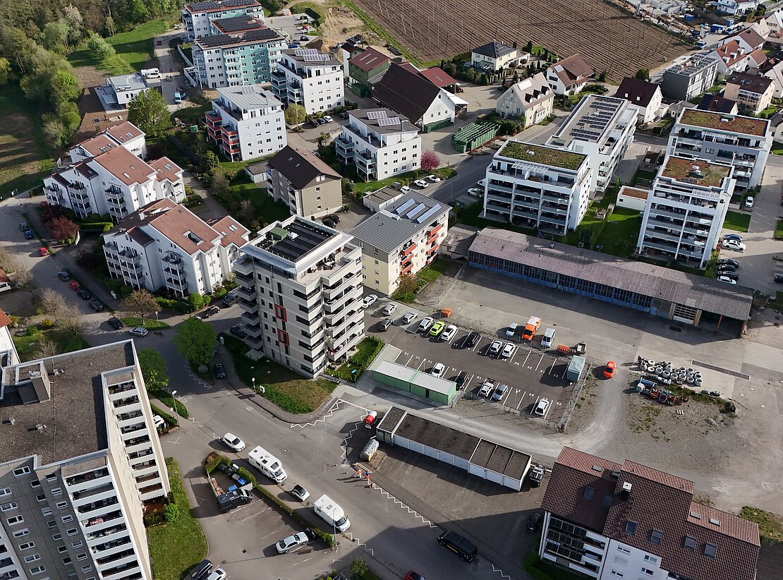 Luftaufnahme eines Wohngebiets mit mehrgeschossigen Wohnhäusern, begrünten Dächern, Solaranlagen und einem Parkplatz mit mehreren Fahrzeugen.