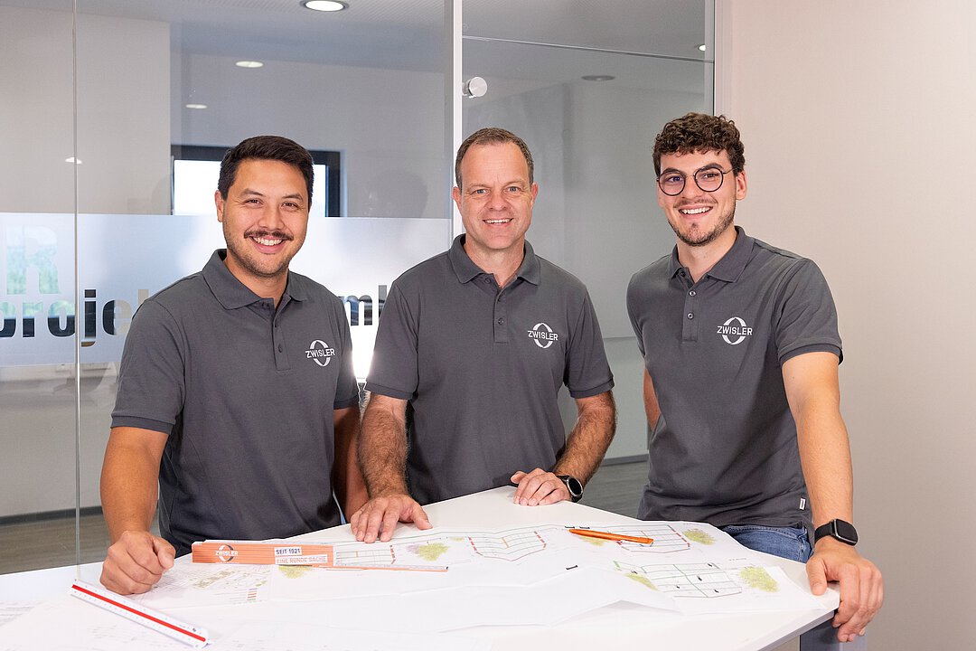 Drei Mitarbeiter der Firma Zwisler Projektbau stehen gemeinsam an einem Tisch mit Bauplänen und lächeln in die Kamera.