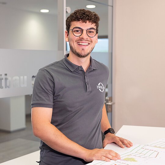 Junger Mitarbeiter von Zwisler Projektbau lächelt in die Kamera, vor ihm liegen Baupläne auf dem Tisch.