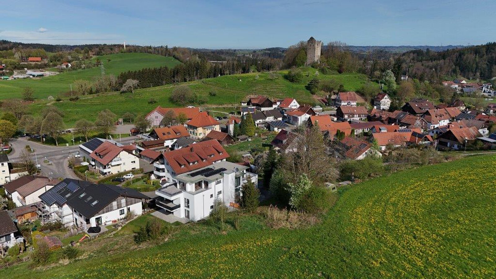 Luftaufnahme von Neuravensburg mit der historischen Burgruine auf einem bewaldeten Hügel im Hintergrund und einem modernen Wohnhaus am Ortsrand.