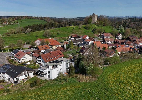 Luftaufnahme von Neuravensburg mit der historischen Burgruine auf einem bewaldeten Hügel im Hintergrund und einem modernen Wohnhaus am Ortsrand.