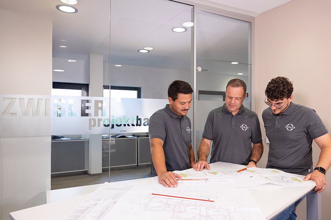 Drei Mitarbeiter von Zwisler Projektbau betrachten gemeinsam Baupläne auf einem Tisch im modernen Büro.