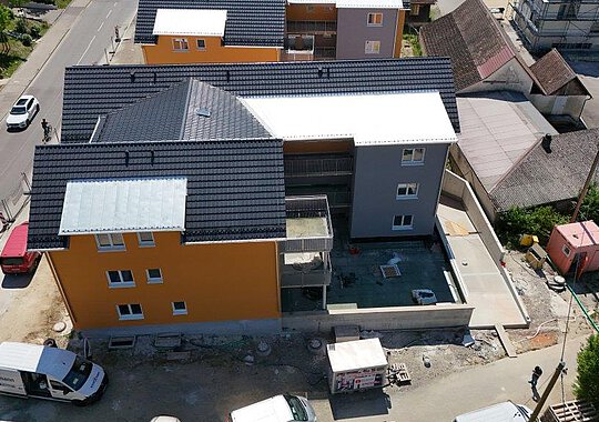 Luftaufnahme eines Wohngebäudes mit Innenhof und Balkonen, teilweise im Rohbauzustand, umgeben von angrenzenden Gebäuden und Baustelleninfrastruktur.