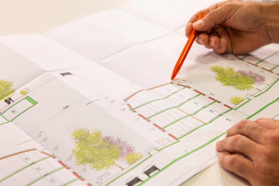 Nahaufnahme einer Hand mit orangefarbenem Stift, die auf einen detaillierten Bauplan mit Grünflächen zeigt.