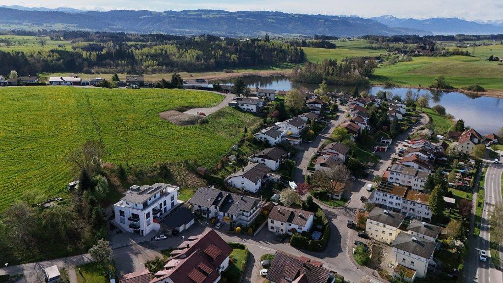 Luftaufnahme von Neuravensburg mit der historischen Burgruine auf einem bewaldeten Hügel im Hintergrund und einem modernen Wohnhaus am Ortsrand.